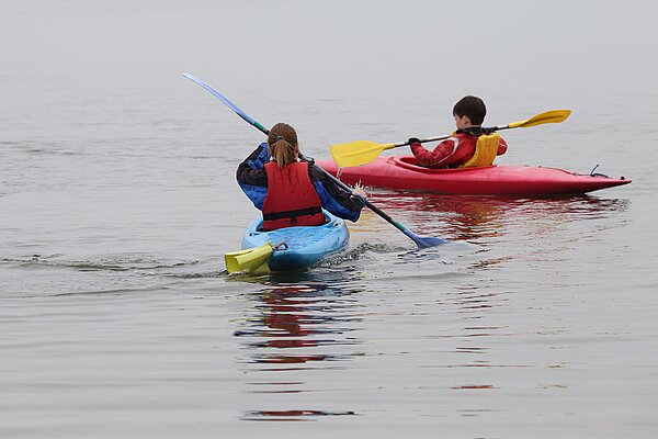 Two children kayaking away from the camera, a girl in a blue canoe, and a boy in a red one, photo is from 2017 when the original centre was up and running. 