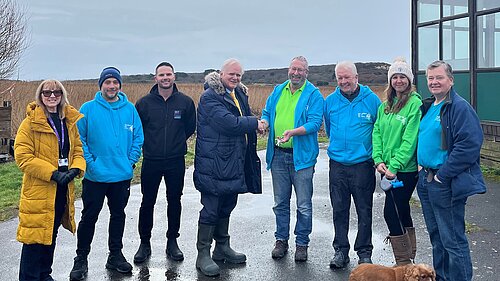 Members of Hengistbury Head Outdoors with Councillor Mike Cox and the BCP Council team posing in a line outside the centre. There are two women and six men in the group, and a brown spaniel. It's a grey, wet day and five of the people are wearing matching brightly-coloured blue or green hoodies